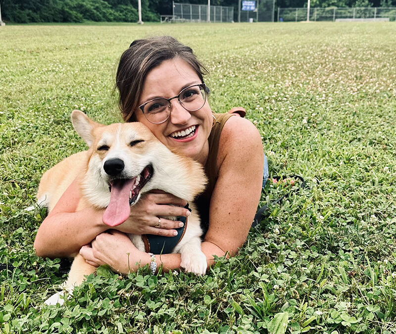 A person with glasses lies on grass, hugging a happy corgi with its tongue out in an open field.