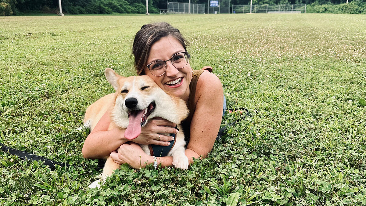 A person with glasses lies on grass, hugging a happy corgi with its tongue out in an open field.