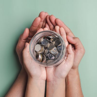 Several hands of different sizes hold a glass jar filled with coins against a green background.