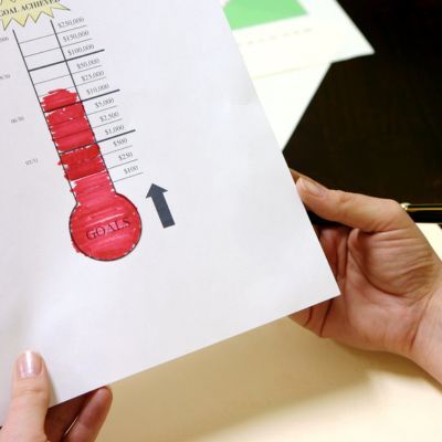 Hands holding a paper with a goal achievement thermometer chart, nearly fully colored in red, indicating close to reaching the target amount.