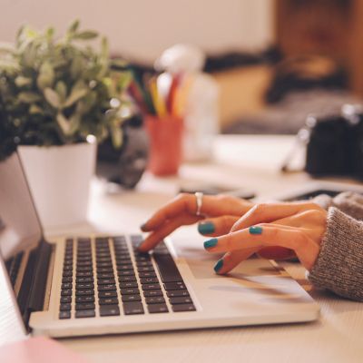 Hands with teal nail polish typing on a laptop keyboard, with a potted plant and colorful pens in the background.