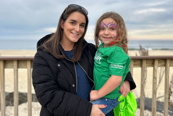 A woman wearing a black coat and sunglasses on her head holds a young girl with face paint and a green shirt, standing in front of a wooden fence by the beach.