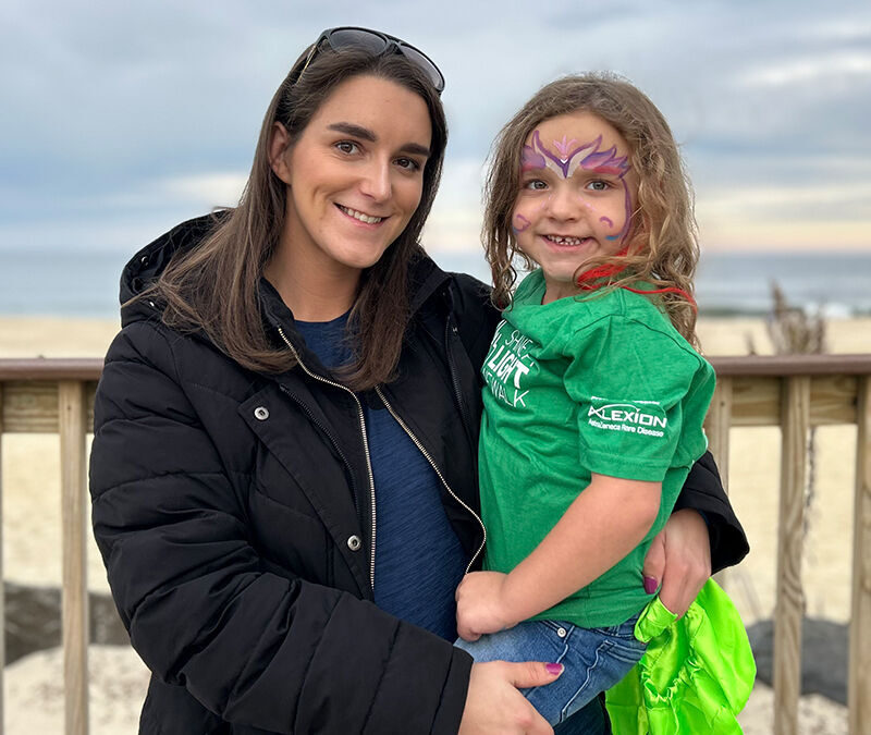 A woman wearing a black coat and sunglasses on her head holds a young girl with face paint and a green shirt, standing in front of a wooden fence by the beach.