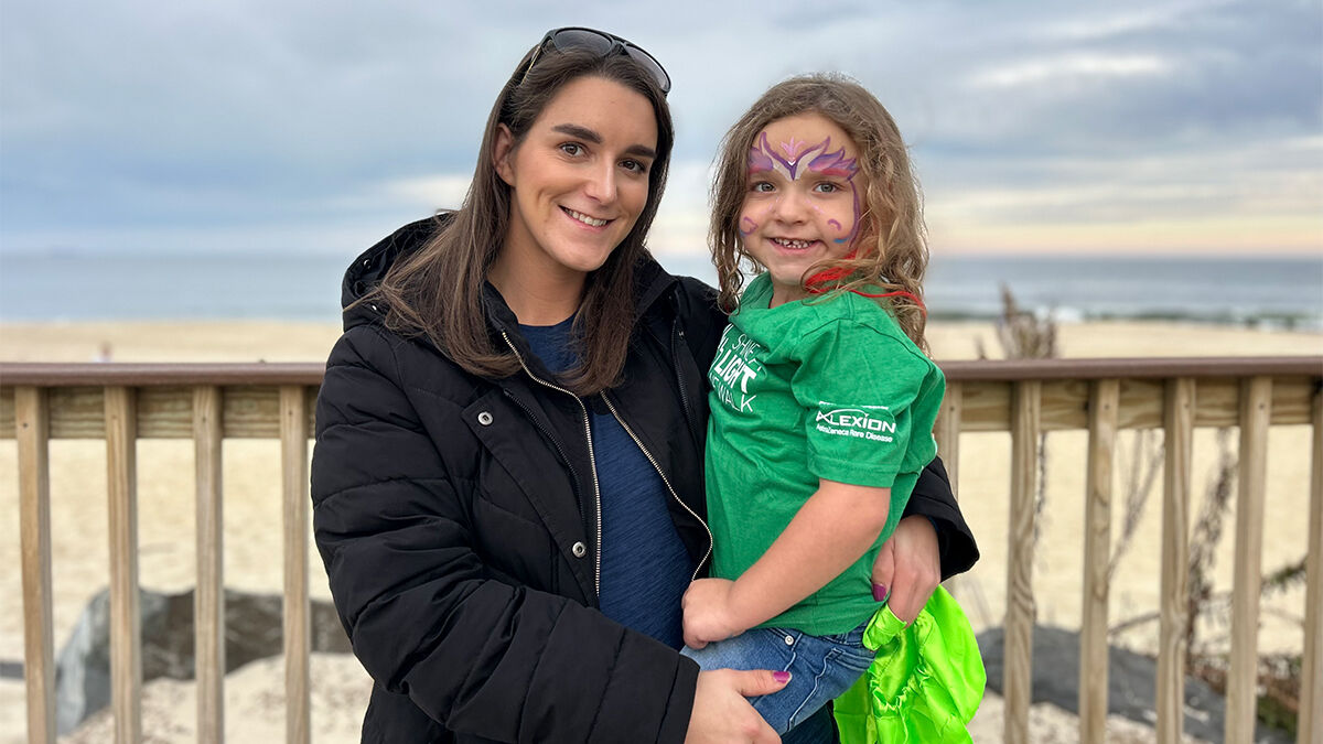 A woman wearing a black coat and sunglasses on her head holds a young girl with face paint and a green shirt, standing in front of a wooden fence by the beach.