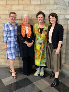 Four women standing side by side in front of a stone wall, smiling at the camera. They are wearing a variety of colorful and formal outfits.