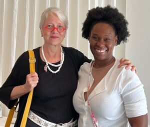 Two women stand next to each other, smiling. The woman on the left has short gray hair and red glasses, and the woman on the right has curly black hair. Both have an arm around the other's shoulder.