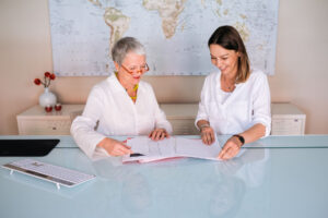 Two women in an office review documents at a glass desk with a world map in the background. Both are smiling and appear engaged in the discussion.