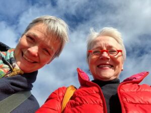 Two people wearing jackets and scarves smile at the camera against a backdrop of a partly cloudy sky.