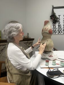 An elderly woman with grey hair works on painting a sculpted bust figure on a table in an art studio, with paint jars, brushes, and glasses nearby. A wall calendar is seen in the background.