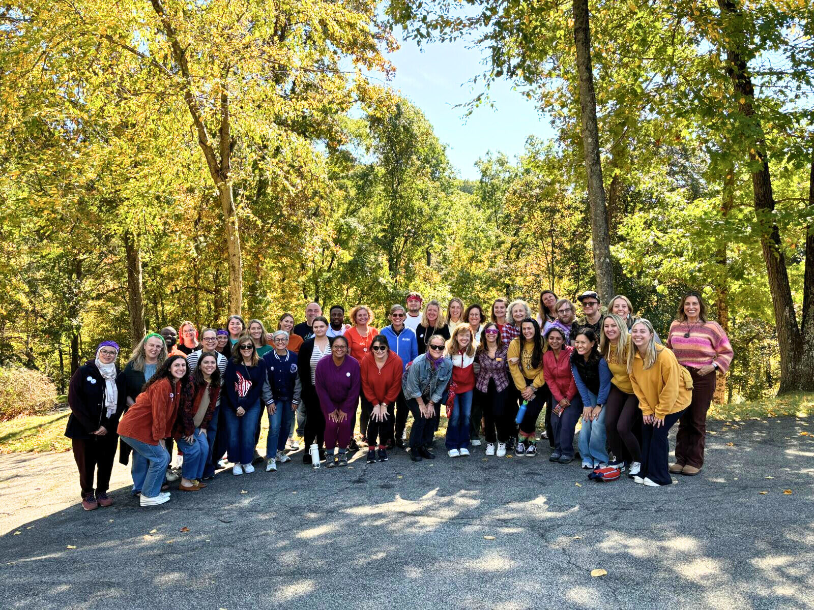 A large group of people posing together outdoors on a sunny day, surrounded by trees with green and yellow leaves.