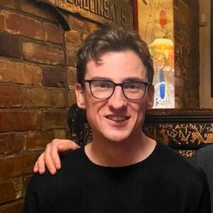 A person with glasses smiles at the camera, standing against a brick wall with posters in the background.