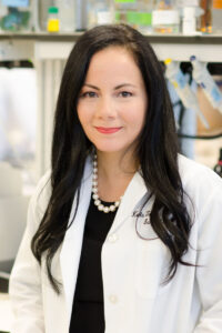 A woman with long black hair wearing a lab coat and a pearl necklace stands in a laboratory.