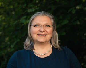 A smiling woman with glasses, gray hair, and a necklace stands in front of green foliage.