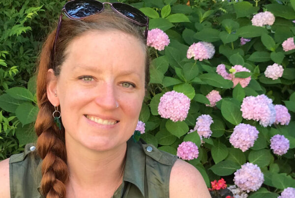 A person with a braid and sunglasses on their head smiles in front of pink hydrangea flowers and greenery.