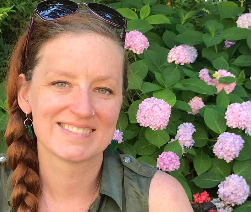 A person with a braid and sunglasses on their head smiles in front of pink hydrangea flowers and greenery.