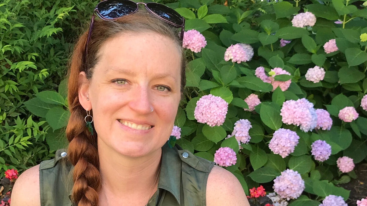 A person with a braid and sunglasses on their head smiles in front of pink hydrangea flowers and greenery.