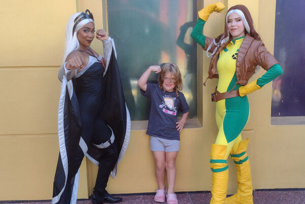 Two people in superhero costumes and a child pose with flexed arms in front of a building.