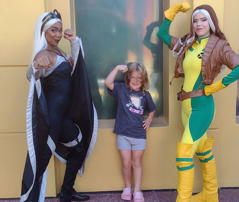 Two people in superhero costumes and a child pose with flexed arms in front of a building.