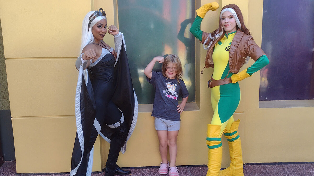 Two people in superhero costumes and a child pose with flexed arms in front of a building.