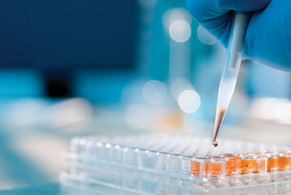 A gloved hand uses a pipette to add liquid to a multiwell plate in a laboratory setting.