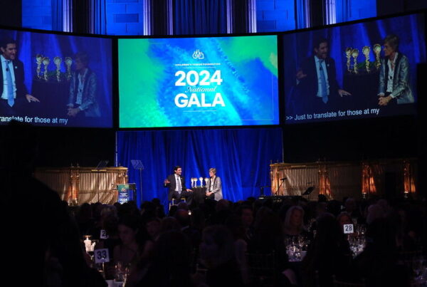 Two people sit on stage with microphones at the 2024 National Gala. Large screens display the event name in a dimly lit venue filled with attendees.