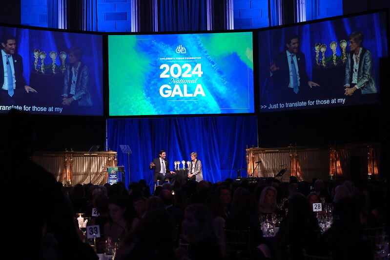 Two people sit on stage with microphones at the 2024 National Gala. Large screens display the event name in a dimly lit venue filled with attendees.