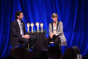 Two people seated and talking on stage against a blue curtain backdrop, with trophies on a table between them.