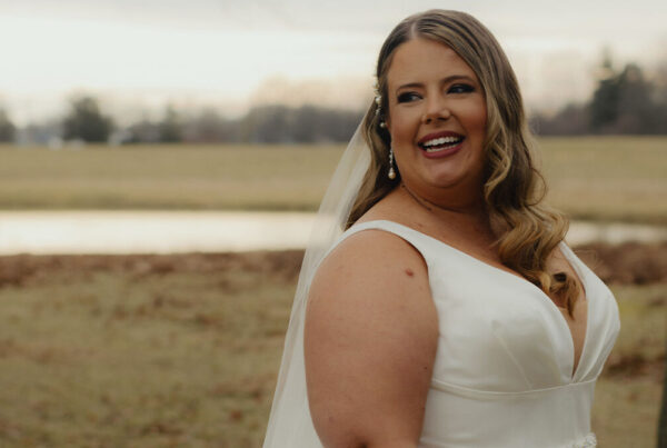 A bride in a white dress and veil stands outdoors, smiling, with a pond and trees in the background.