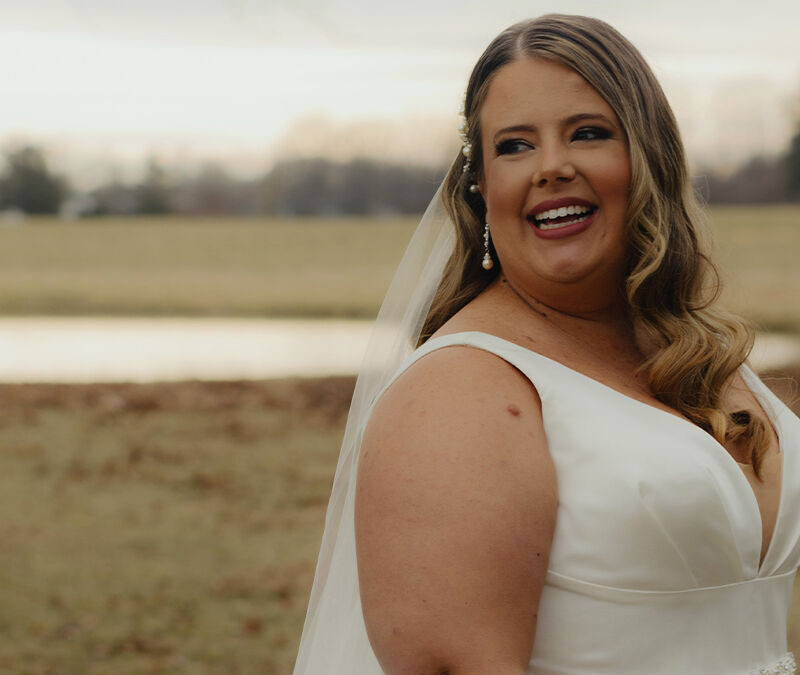 A bride in a white dress and veil stands outdoors, smiling, with a pond and trees in the background.