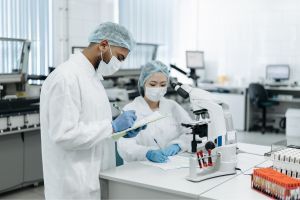 Two scientists in lab coats and masks work near a microscope in a laboratory setting.