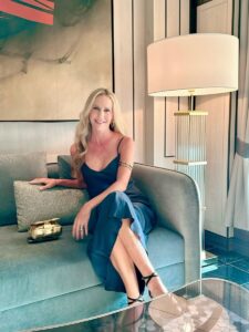 A woman in a dark dress sits on a sofa next to a lamp, holding a cushion. A clutch bag rests beside her.