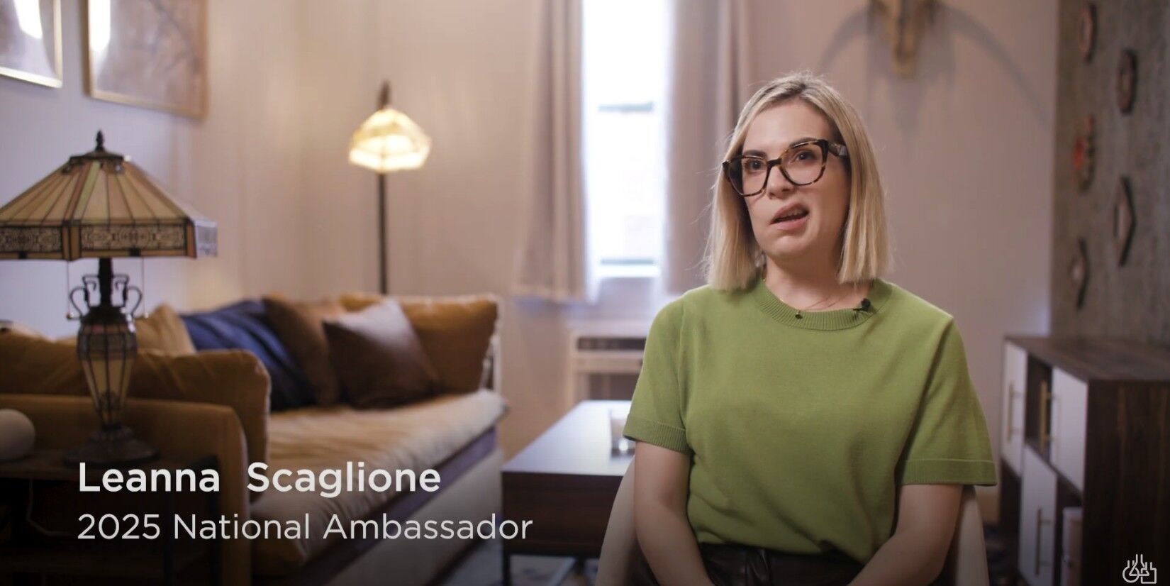 A person in a green shirt sits in a living room with lamps and a sofa in the background. Text on the image reads "Leanna Scaglione, 2025 National Ambassador.