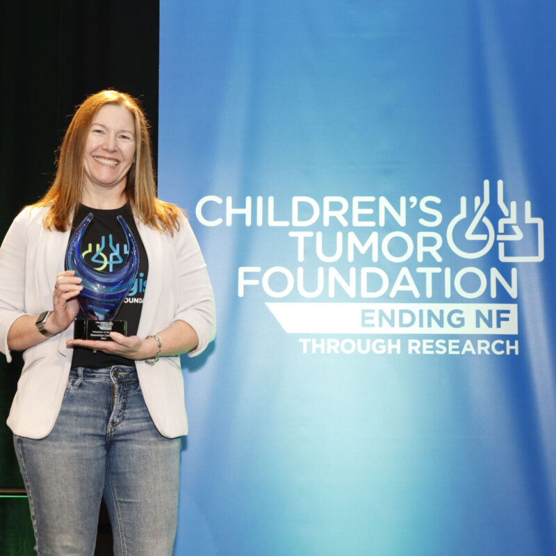 A woman holding an award stands beside a blue banner that reads, "Children's Tumor Foundation: Ending NF Through Research.