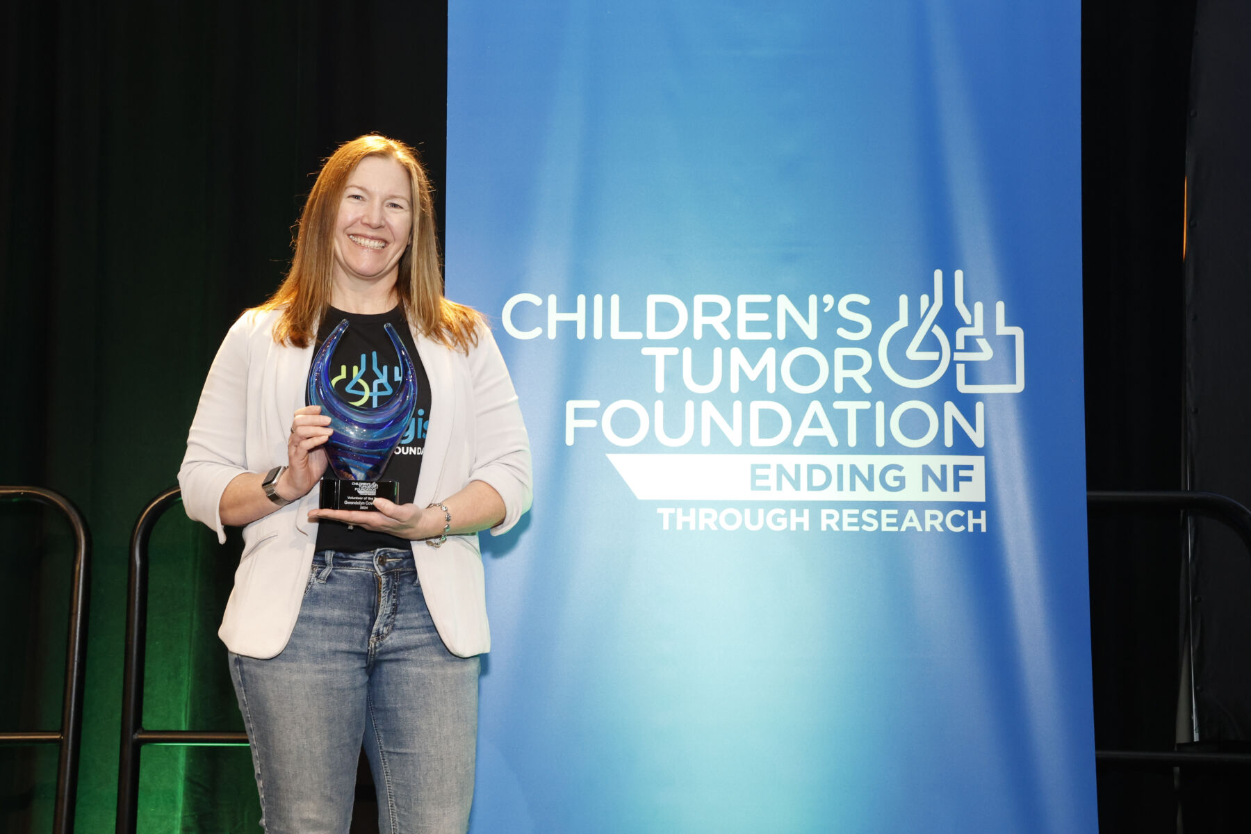 A woman holding an award stands beside a blue banner that reads, "Children's Tumor Foundation: Ending NF Through Research.