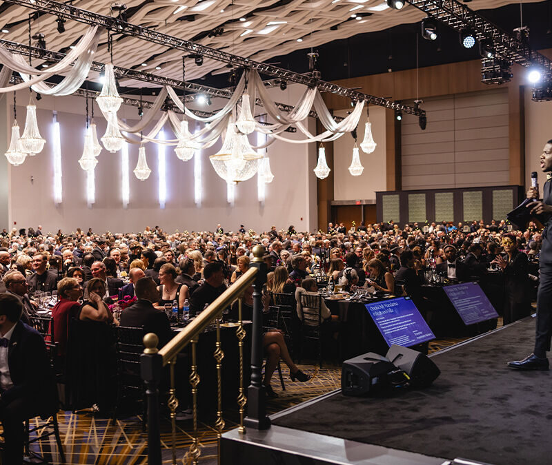 A crowd seated in a large banquet hall listens to speakers on stage, with chandeliers and decorative drapery overhead.