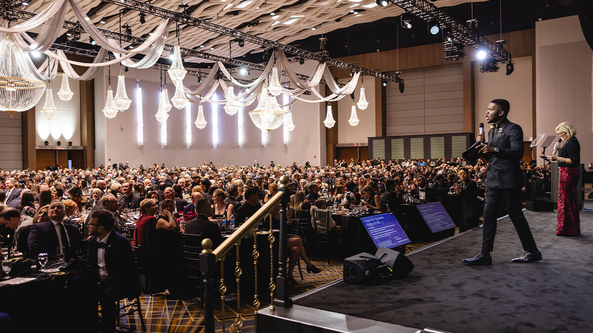 A crowd seated in a large banquet hall listens to speakers on stage, with chandeliers and decorative drapery overhead.