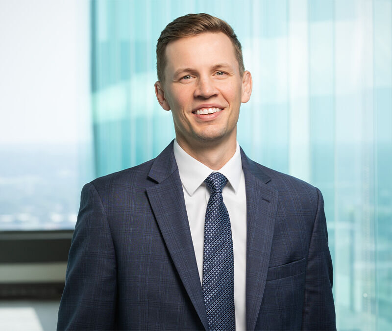 A smiling person in a suit stands in front of tall glass windows with a cityscape in the background.