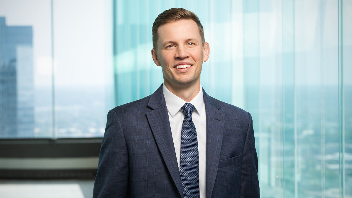 A smiling person in a suit stands in front of tall glass windows with a cityscape in the background.