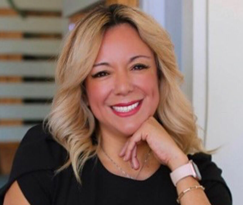 A woman with blonde hair smiles and poses with her hand on her chin, wearing a black top and a smartwatch, in an indoor setting.