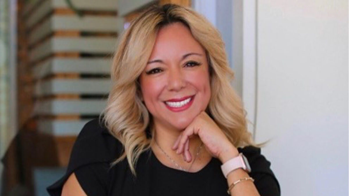 A woman with blonde hair smiles and poses with her hand on her chin, wearing a black top and a smartwatch, in an indoor setting.