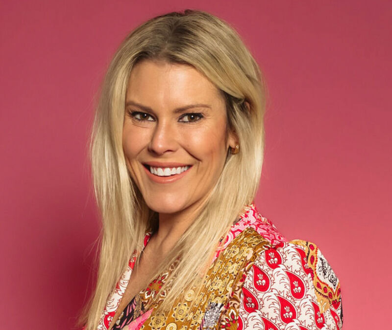 A person with long blonde hair smiles against a pink background, wearing a colorful patterned blouse.