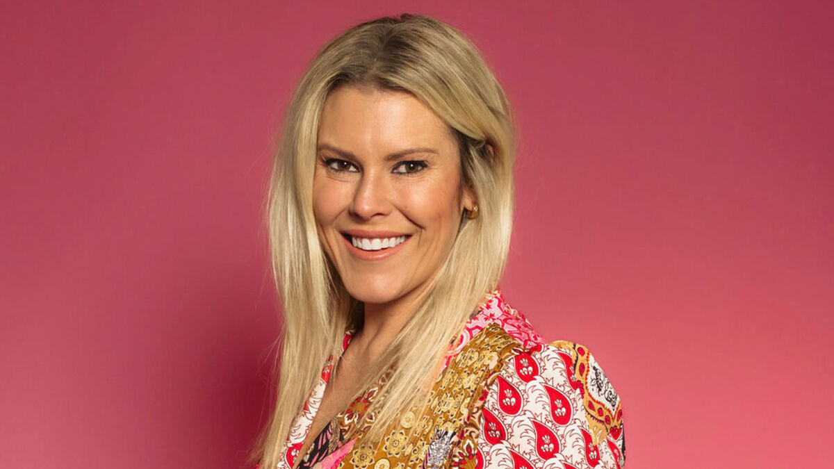 A person with long blonde hair smiles against a pink background, wearing a colorful patterned blouse.