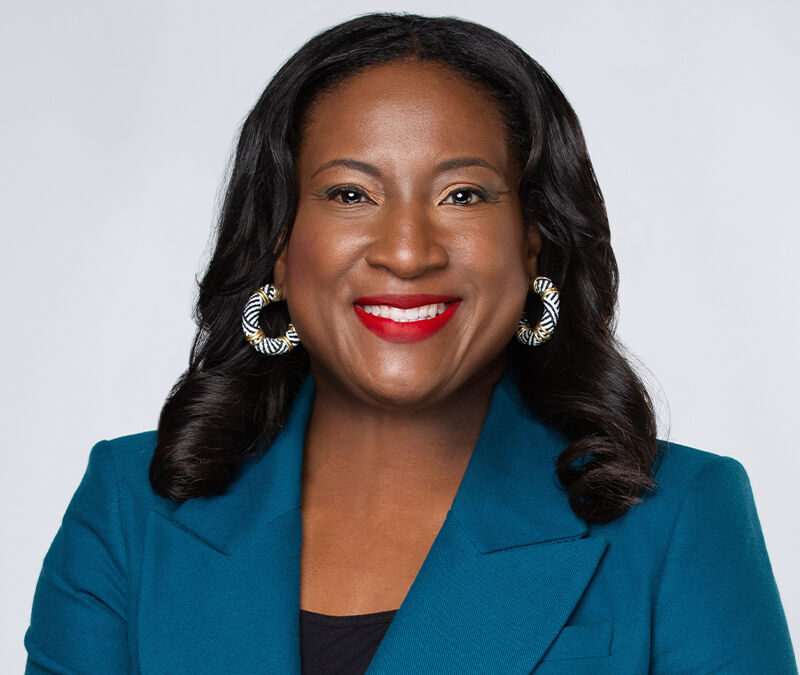 A woman with long dark hair, wearing a teal blazer, statement earrings, and bright red lipstick, smiles against a plain background.