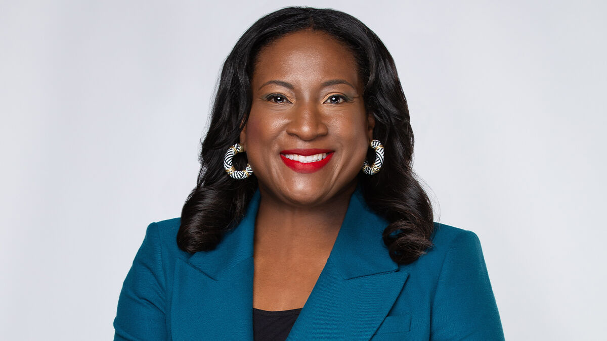 A woman with long dark hair, wearing a teal blazer, statement earrings, and bright red lipstick, smiles against a plain background.