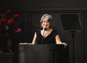 An older woman with gray hair speaks at a podium with two microphones. She is wearing glasses and a black dress. There are flowers in the background.