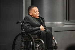 A man in a wheelchair wearing a formal black suit smiles warmly.