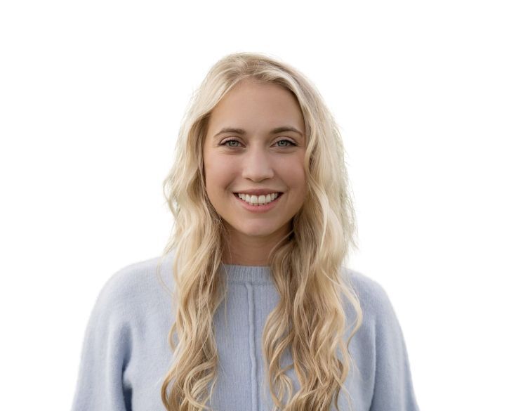 A person with long blonde hair, wearing a light blue sweater, smiles at the camera against a plain white background.