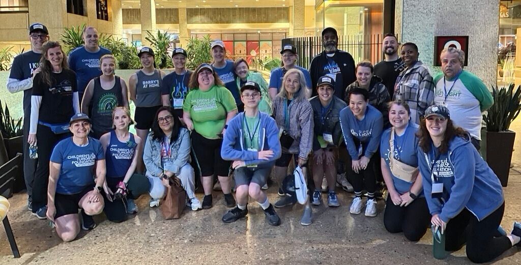 A diverse group of people gather indoors for a group photo, some wearing event-themed shirts and hats. They stand and squat in rows, smiling at the camera.