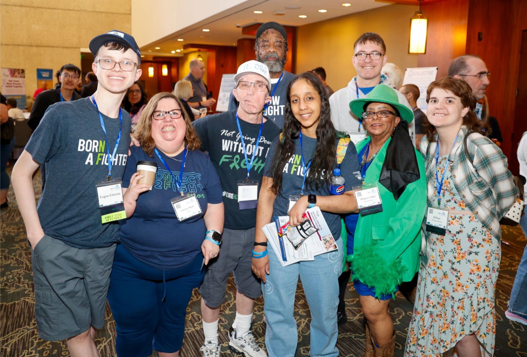 A group of people wearing event badges stands together, smiling. Some are wearing T-shirts with slogans and holding programs. The setting appears to be a conference or gathering.