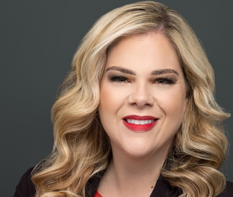 A person with long wavy blonde hair, wearing a black blazer and a red top, smiling in front of a plain dark background.